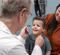 Pediatrician uses an otoscope to examine a smiling young boy's ear while his mother watches.